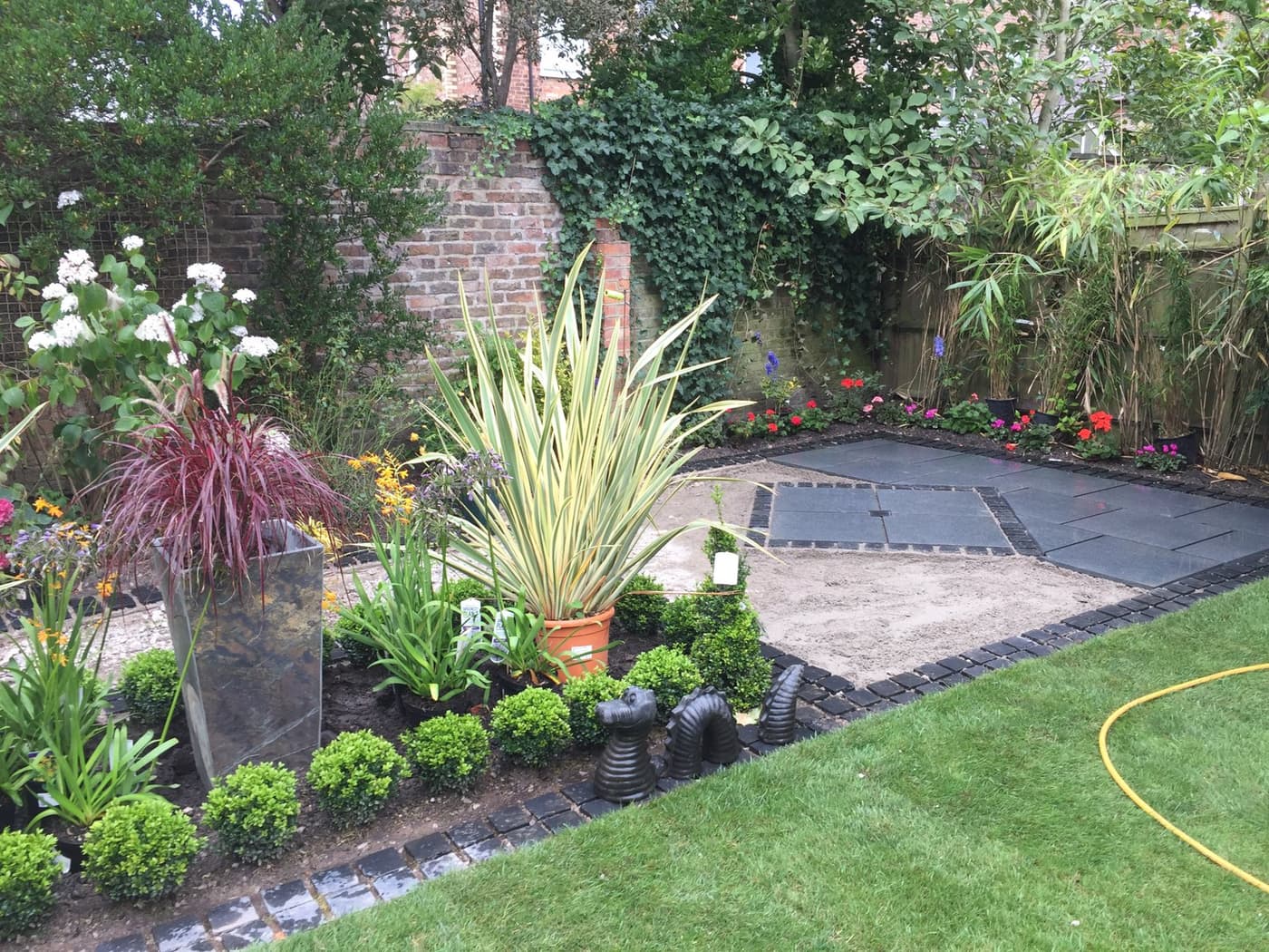 New granite patio under construction, bordered by mixed planting including phormiums, box balls and bamboo screening