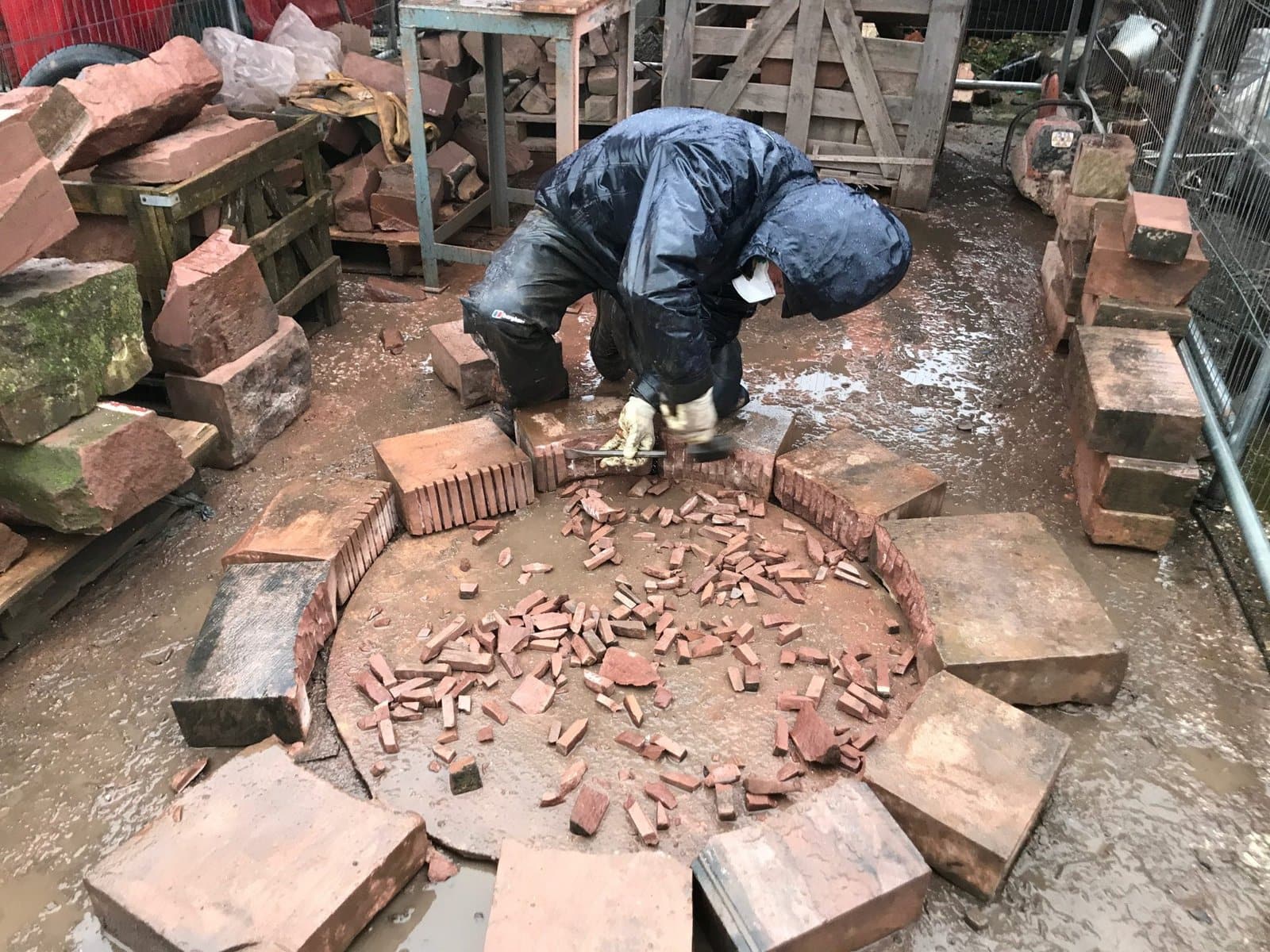 Jason Costello hand-cutting reclaimed brick for a circular stone feature, working through the rain